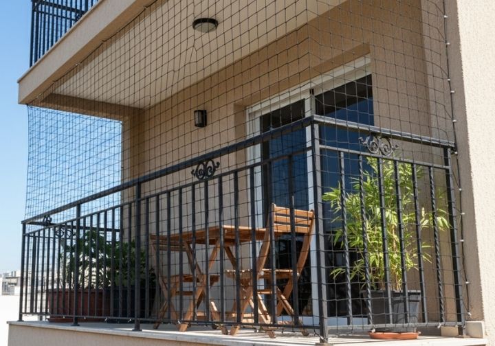 Varanda de apartamento protegida por uma rede de segurança de polietileno preta, com mesa de madeira e plantas.