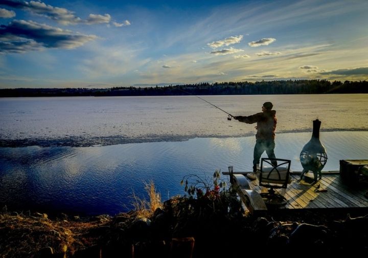 A man fishing in a lake at sunset, with golden reflections in the water and silhouettes of trees in the background.