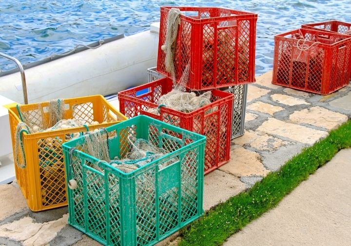 Caixas de plástico vermelhas, verdes, amarelas e cinzentas dispostas num cais junto à água, contendo redes de pesca enroladas e boias.