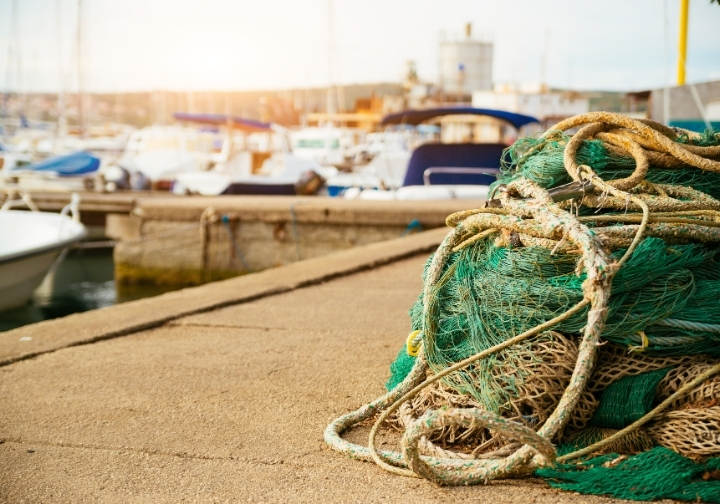 Green fishing nets and thick beige ropes piled on a concrete dock, with blurred white boats and sunlight in the background of a harbour.