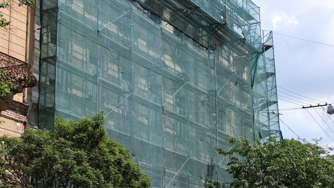 Fachada de edifício em reabilitação totalmente coberta por andaimes metálicos e rede de proteção verde.